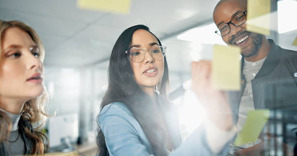 istock Woman, writing and sticky note with teamwork, glass board for project ideas and agenda schedule. Collaboration, problem solving and group in business discussion for brainstorming meeting in office 2225766039