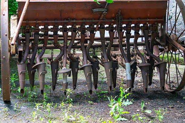 old rusty seed drill with metal funnels and seeding mechanisms, surrounded by weeds.