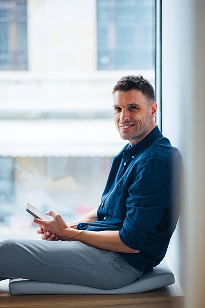 istock Smiling Man Sitting by a Window Holding a Smartphone in Relaxed Pose 2225770901