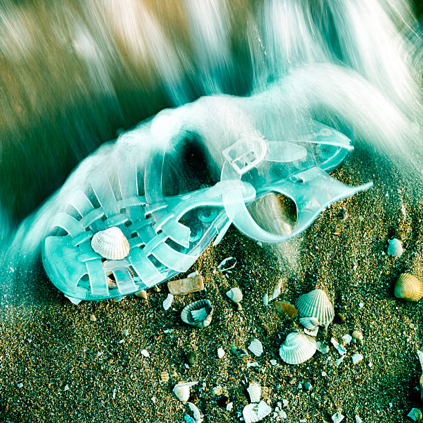A child's plastic sandal lies abandoned on a shell-covered beach, touched by foamy waves — a silent trace of displacement, evoking themes of migration, loss, and fragile innocence.