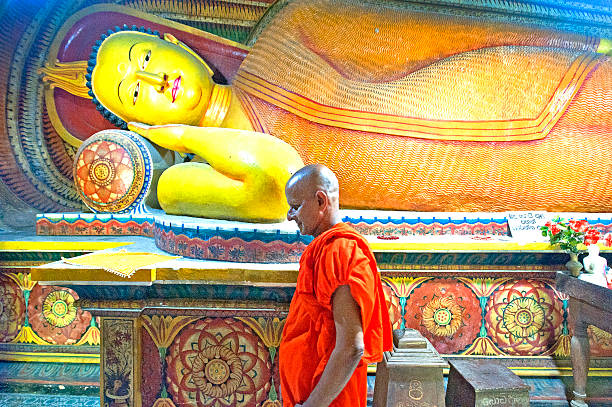 Old Buddhist monk walks past reclining buddha, Samudragiri Viharaya Buddhist temple, Mirissa, Sri Lanka. Samudragiri Viharaya, aka Veheragalla Samudragiri Viharaya, Buddhist temple renowned for its well-preserved murals from the Kandyan era, showing Jataka tales and Buddhist themes. Sri Lanka is the tropical Indian Ocean island formerly known as Ceylon, situated off the southeastern tip of India, renowned for its tea, rubber and precious stone production throughout the world.