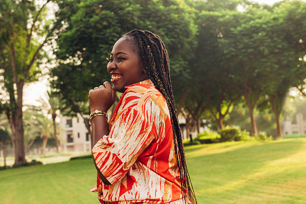 A woman smiles brightly as she stands in the soft shade of tall trees. Dressed in a vibrant orange patterned outfit, her joyful energy shines through as her long braided hair flows down her back. The lush green surroundings provide a serene backdrop, emphasizing the lightness and freedom of the moment. She radiates pure happiness, basking in the peaceful atmosphere of the outdoors.