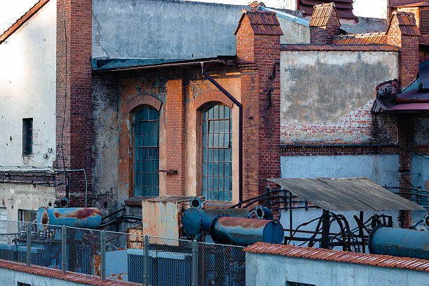 A close-up of a weathered red brick industrial chimney with metal reinforcements, standing next to an old factory building with broken windows. The image captures the decay of an abandoned industrial site, symbolizing urban decline and history.