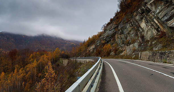 A curving mountain road flanks steep rock faces and traverses a vivid autumn forest. Clouds hover low over rugged peaks, adding a mystical mood to the scene. The contrast between the warm fall colors and the overcast sky provides a striking visual appeal. This image captures the serene beauty of an autumn mountain landscape.