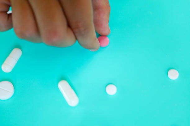 A human hand picks up a pink pill among scattered white tablets and capsules on a bright turquoise background. The image symbolizes the process of taking medication, selecting drugs, or health care.