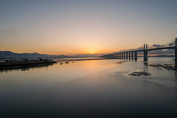 The photo shows the bridge on concrete piers stretching across exposed mudflats. First light hits the  rails; Low tide reveals channels and small pools beside the tracks. Use: coastal transport, sunrise rail imagery, engineering stock.