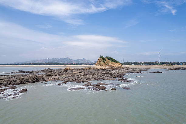 The photo captures a flat, rocky island ringed by light-colored water. Sparse grass and a single cluster of low bushes occupy the center; the surrounding sea shows gentle ripples. No buildings or boats appear. Uses: island geography, marine travel, nature documentation, aerial seascape, remote location imagery.