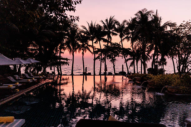 A breathtaking pink and orange sunset over a tropical landscape in Thailand, with silhouettes of palm trees and their reflections shimmering in a serene swimming pool.