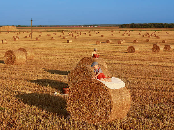 Children in vintage clothes playing and resting on hay bales in a wide harvested field. Rural childhood, warm sunset light, copy space.
