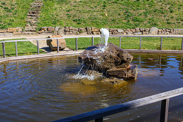 Public park pond with a fountain, water gushing from water spout over huge stone, surrounded by railing, village street in background, sunny day in Gulpen, South Limburg, Netherlands