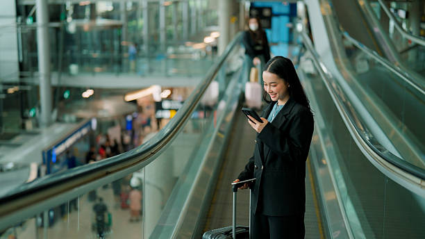 istock Young Asian businesswoman using mobile phone and smiling while going up a moving walkway in an airport. 2225822716