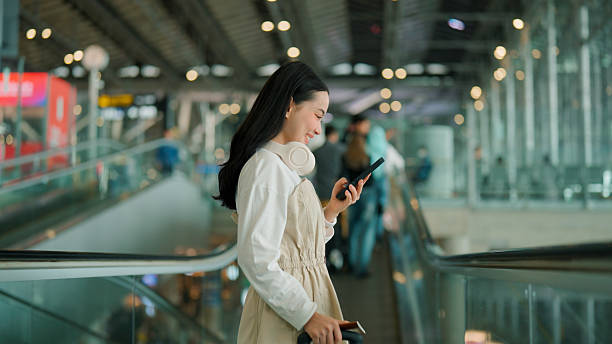 istock Young Asian woman using mobile phone and smiling while going to the upper floor on a moving walkway in an airport. 2225824259