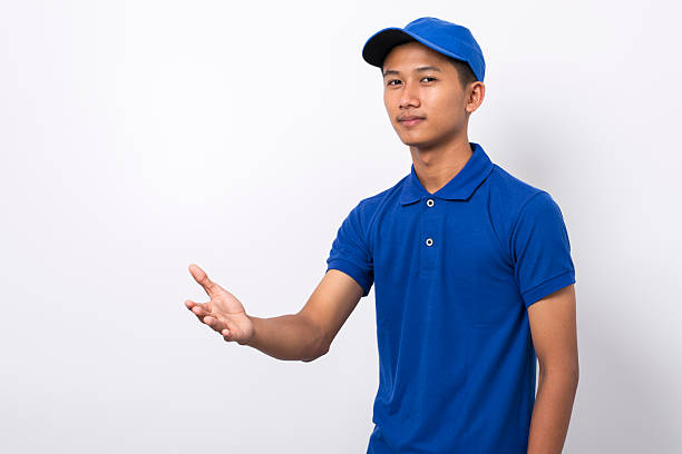 istock Smiling young asian delivery man wearing blue uniform pointing present a product 2225825317
