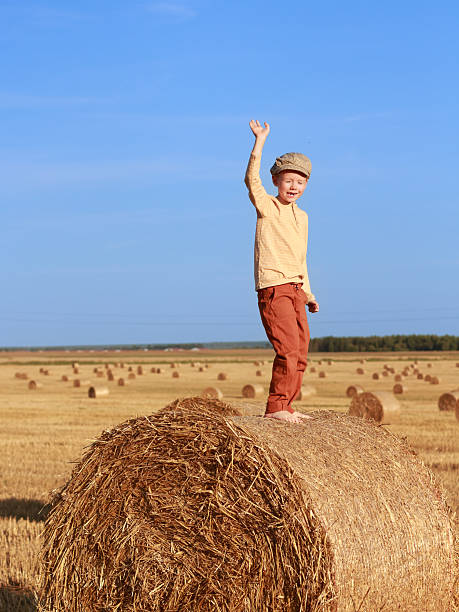 Boy in retro outfit stands barefoot on a large hay bale, raising his hand in greeting. Wide countryside field, clear blue sky.
