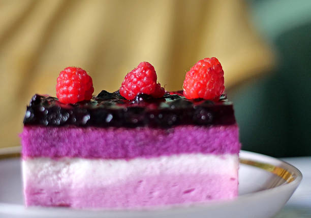 A captivating close-up showcases a single, perfectly cut slice of multi-layered mousse cake, featuring rich purple and soft pink layers, topped with a glistening dark berry glaze and fresh red raspberries. This decadent dessert piece highlights intricate patisserie artistry, ideal for themes of gourmet food, sweet treats, celebrations, or high-quality bakery offerings.