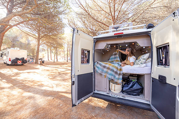 Woman lying on bed inside camper van with cozy blanket and pillows. Outdoor camping scene with trees and RVs in background. Van life lifestyle and nature relaxation concept.