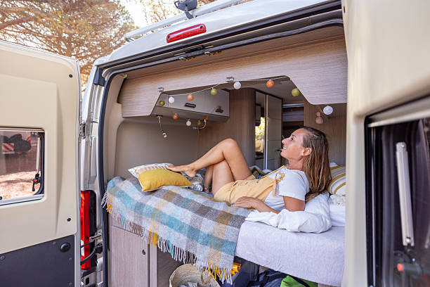 Young woman lying on a bed inside a camper van with a plaid blanket and pillows. Comfortable van life interior with storage and soft lighting. Casual lifestyle and travel living space.