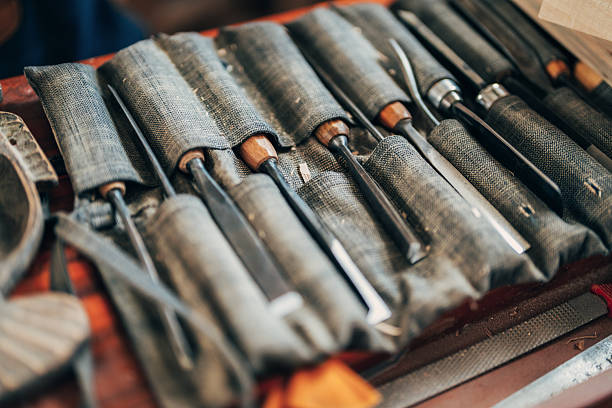 This photo shows a neatly arranged set of woodworking chisels and carving tools. The tools are organized in a fabric roll-up pouch, with wooden handles peeking out of their compartments. The pouch lies open on a wooden workbench, revealing a variety of tools used for fine craftsmanship, likely in wood carving or detailed joinery.
