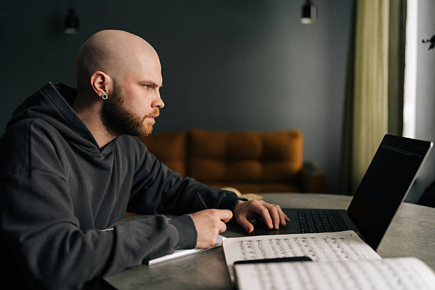 istock Side view of talented composer writing music notes on paper sitting at table with laptop, books, sheet music in home office, immersing in creativity and inspiration. 2225889292