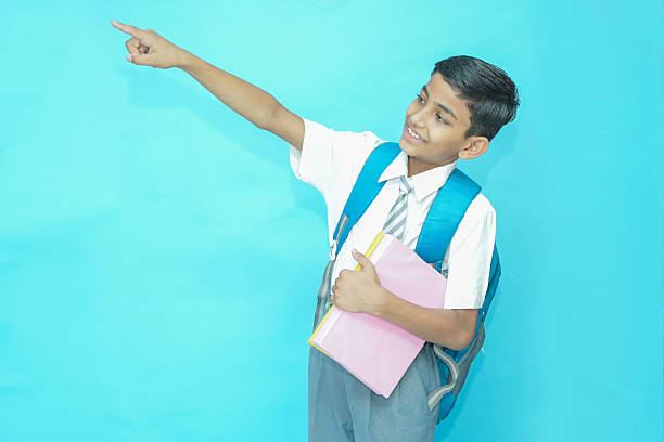 Indian student schoolboy wearing uniform holding notebooks and bag pointing at blank space isolated over blue background, Education knowledge concept. Look here. Admission open.