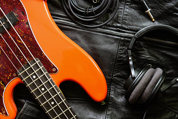 Close-up focus on a guy's hand clenching a chord on the neck of a guitar. A man is playing guitar in a field. The guitar is a vintage model with a black strap. The musician is wearing a white jacket and enjoying playing the guitar
