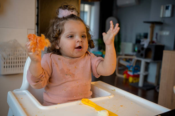 Joyful toddler girl with curly hair raises her hands and smiles while holding fruit during mealtime in a home kitchen. Her pink shirt and expressive gestures add charm to this candid moment of childhood.