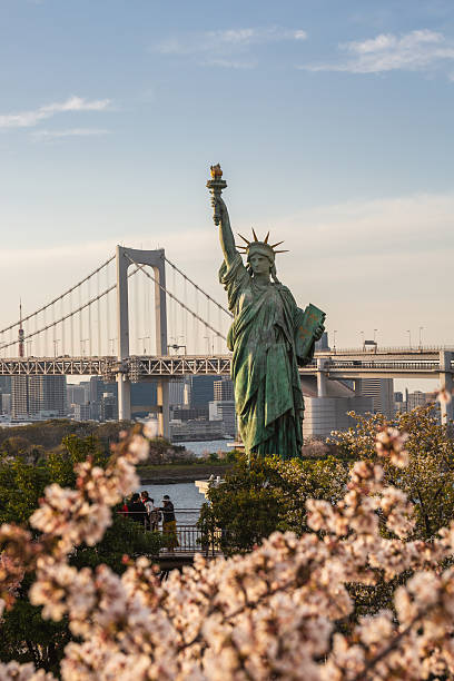 Tokyo, Japan - March 31 2023: Statue of Liberty replica in Odaiba, Tokyo, with Rainbow Bridge and cherry blossoms in the foreground. A captivating view of iconic landmarks.