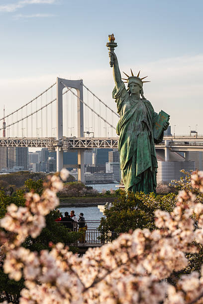 Tokyo, Japan - March 31 2023: Statue of Liberty replica in Odaiba, Tokyo, with Rainbow Bridge and cherry blossoms in the foreground. Tourists enjoying the view.