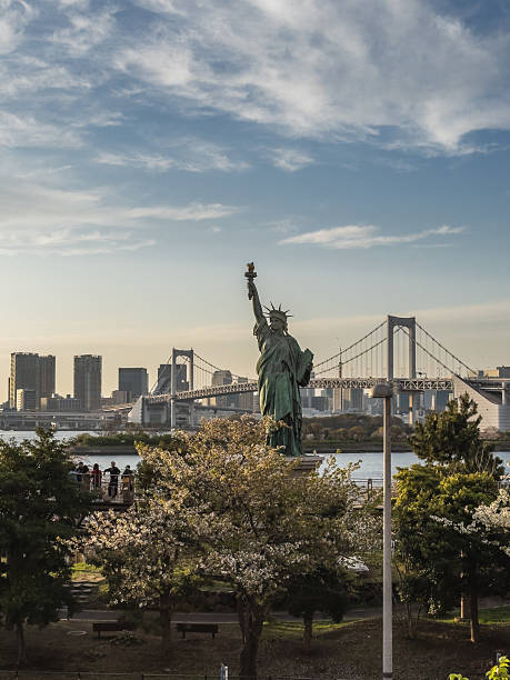 Tokyo, Japan - March 31 2023: Statue of Liberty replica in Odaiba, Tokyo, with Rainbow Bridge and cityscape in the background. Spring cherry blossoms add beauty to the scene.