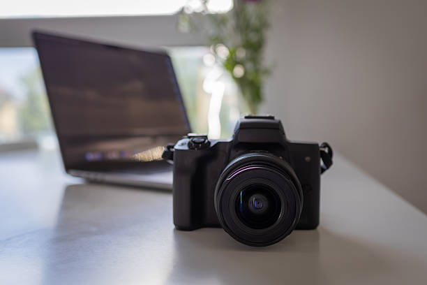 istock Modern digital camera with a prominent lens on a clean white desk, showcasing professional photography equipment and creative digital content creation tools with a laptop 2225940283