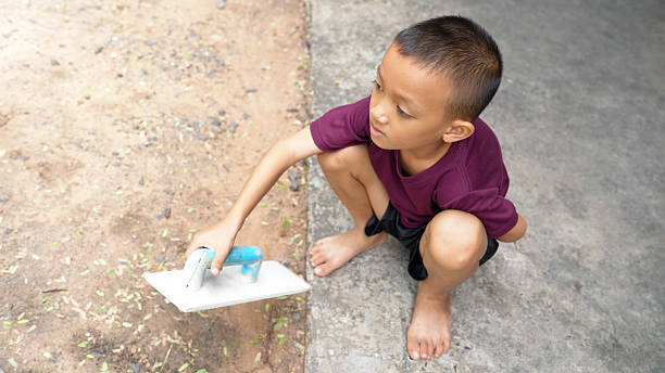 Young boy is kneeling ground, focused using small tool to smooth surface. He is wearing maroon shirt and black shorts, with bare feet. scene captures moment of concentration and playfulness