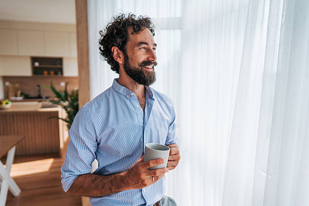 istock Smiling man drinking coffee looking out the window at home 2225944483