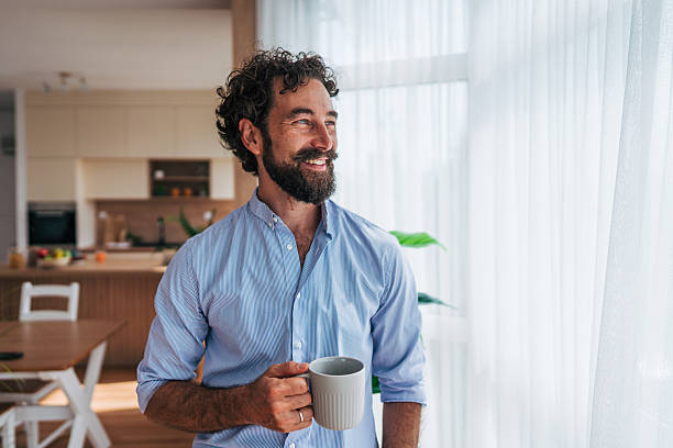 istock Smiling man drinking coffee and looking out the window at home 2225944493