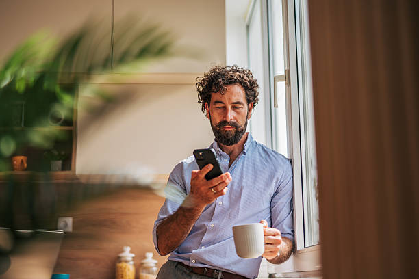 istock Mid adult man using smartphone and drinking coffee in kitchen 2225944625