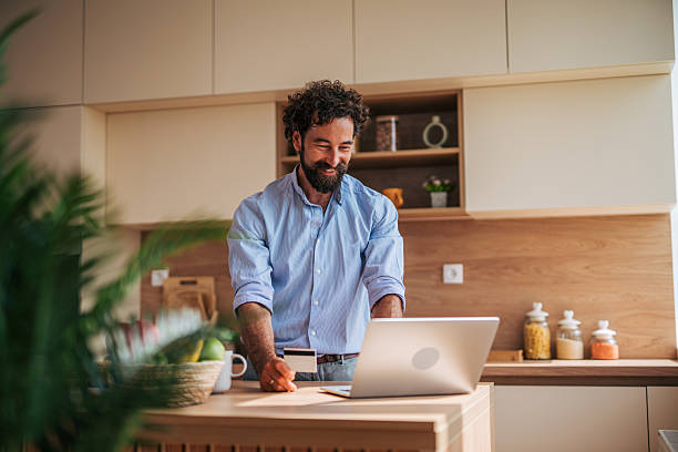 istock Smiling man using laptop and credit card in kitchen 2225945006