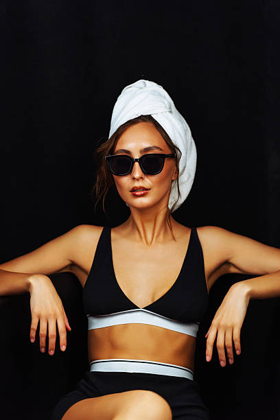 A model relaxes during a skin care treatment, wearing a towel and sunglasses in a chic, dark environment.