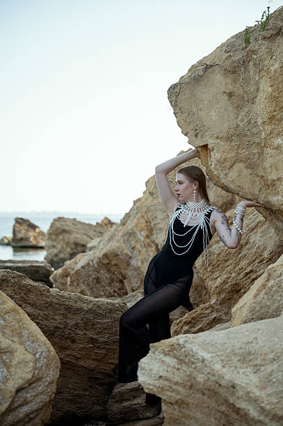 A young blonde poses on a rocky beach in a stylish black suit, with a large pearl necklace on her neck. A person standing in front of a large rock. High quality photo