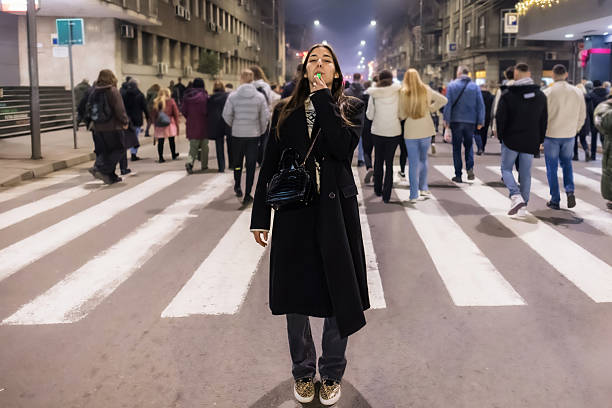 Young woman in warm clothing standing on the street and blowing into whistle during protest in the city
