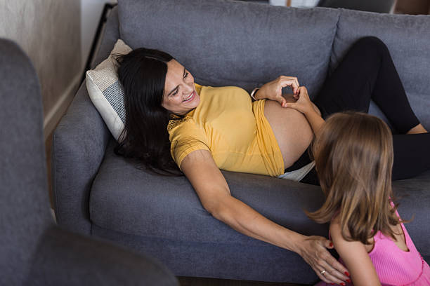Pregnant mother lying on sofa making heart shape on her belly, her daughter looking at her