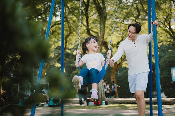 A cheerful young girl enjoys swinging as her father gently pushes her at a playground. Captured during a sunny day, this candid moment reflects happiness, parental support, and childhood fun in nature.
