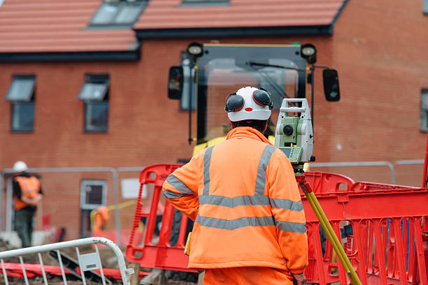 istock Construction worker in bright orange safety gear is using  surveying instrument at  construction site bordered by safety barriers with nearby machinery and workers visible 2226005588