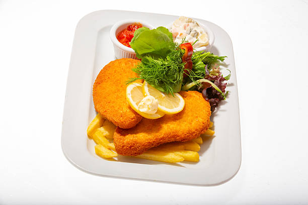 A golden-brown breaded pane cutlet rests on crispy French fries, topped with lemon slices and fresh herbs. Served with creamy coleslaw, tangy tomato dip, and a refreshing side salad, this dish combines crunch, zest, and color in perfect balance.