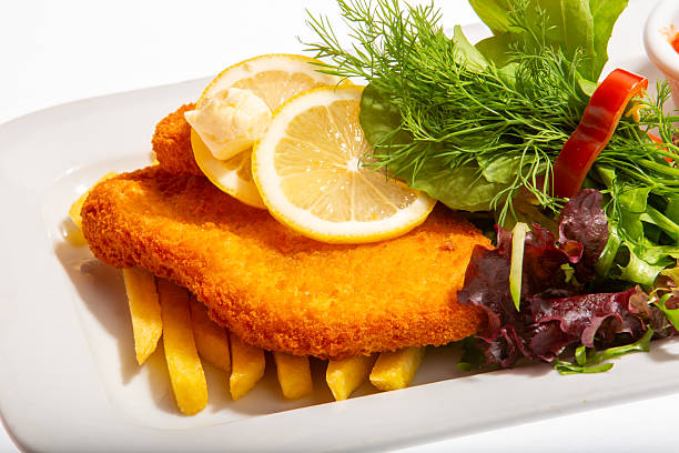 A golden-brown breaded pane cutlet rests on crispy French fries, topped with lemon slices and fresh herbs. Served with creamy coleslaw, tangy tomato dip, and a refreshing side salad, this dish combines crunch, zest, and color in perfect balance.