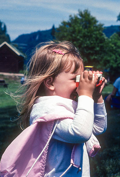 Surprised cute girl 4-5 years old in a straw hat and a yellow t-shirt in the summer stands in a field with tall grass against the blue sky, looks through binoculars. Close up