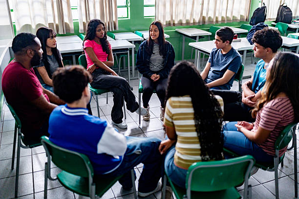 A diverse group of young people is sitting in a circle inside a classroom, participating in a group therapy session. They are engaged in conversation, reflecting a supportive environment focused on mental health and emotional wellbeing