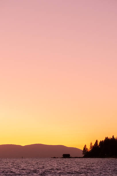 An idyllic scene where a silhouette of a cabin rests, frame-right, on the shore of a mountain lake. The air itself is saturated with the bright orange glow of the setting sun. In the distance silhouetted mountains rise above the water into the cloudless sky where there is a perfect pink to orange gradient.
