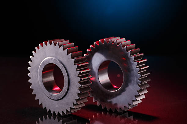 Two metallic gears are positioned closely together on a red surface. The gears are highly polished, reflecting light and creating a glossy appearance. A blue light source is visible in the background