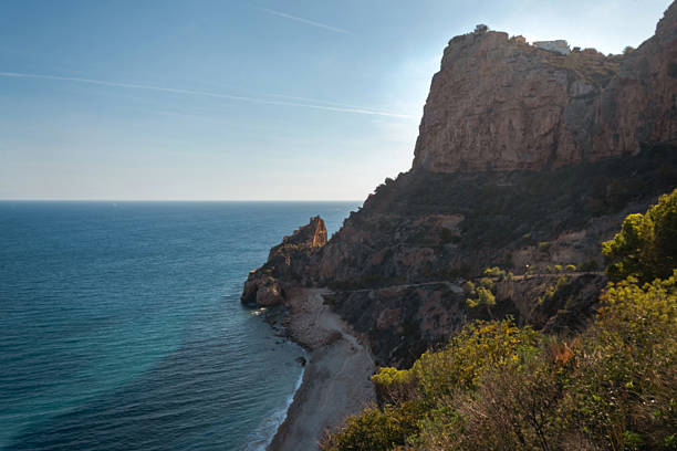 The rock rises above the blue sea in the orange rays of the setting sun against the background of the blue sky