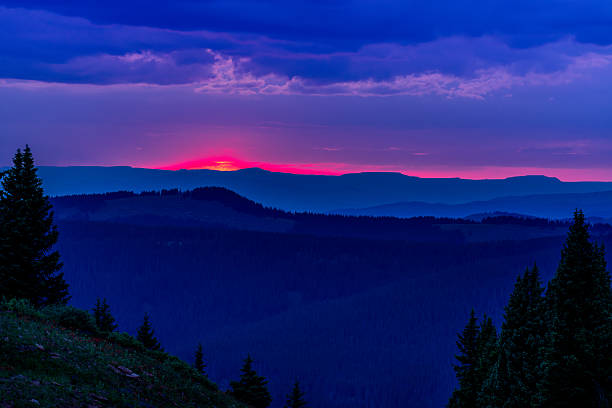 Mountain Sunset with Silhouetted Trees Along Ridge - Scenic summer mountain landscape near Vail, Colorado USA. Sunset peaking through clouds with red light and layers of mountains leading off into the distance.