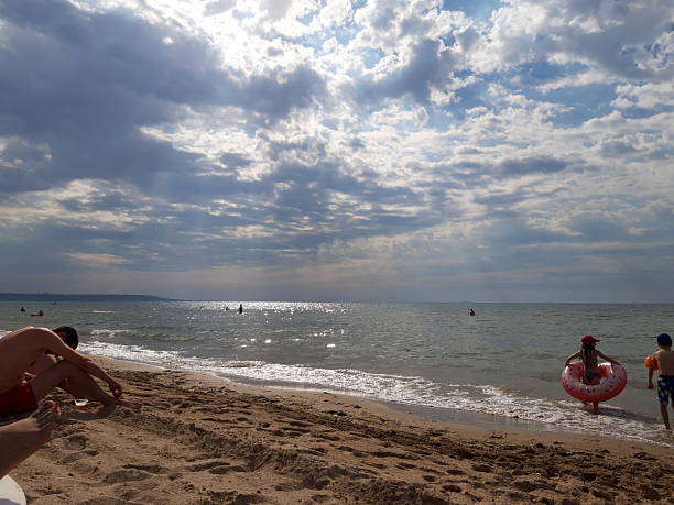 Morning at sea, the sun's rays break through the clouds, two children enter the water, and a man appears to be doing exercises on the sand.